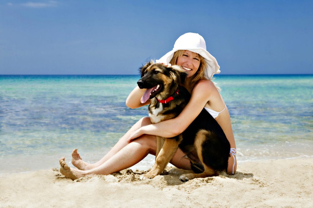 Blond woman sitting on sand hugging her dog.