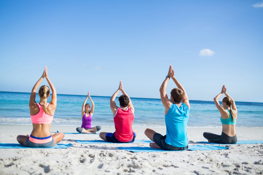 Yogis holding a pose with their hands over their heads while sitting on a beach facing their yoga instructor.