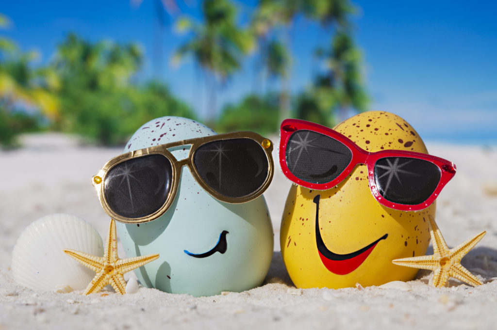 Two Easter eggs with smiles painted on and wearing sunglasses sitting on a beach with a shell and two starfish.