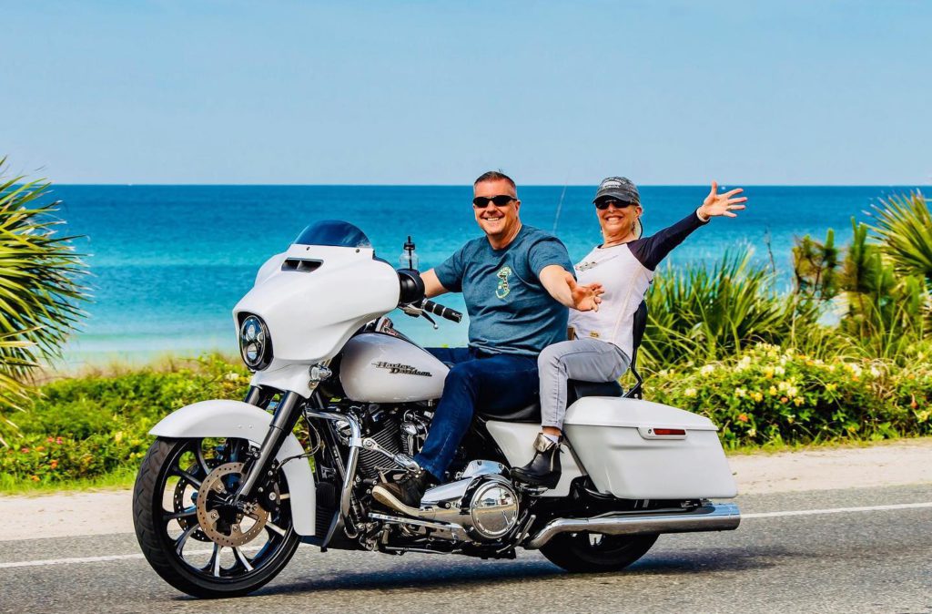 Couple on a Harley Davidson motorcycle waving as they ride alongside the beach. 